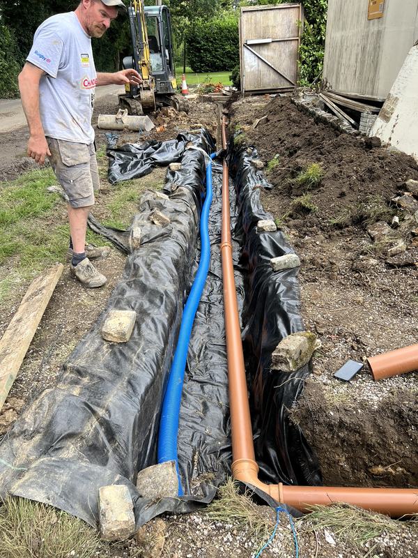 Membrane-lined drainage trench with orange underground drainage pipe and blue duct laid, with a worker and mini digger behind