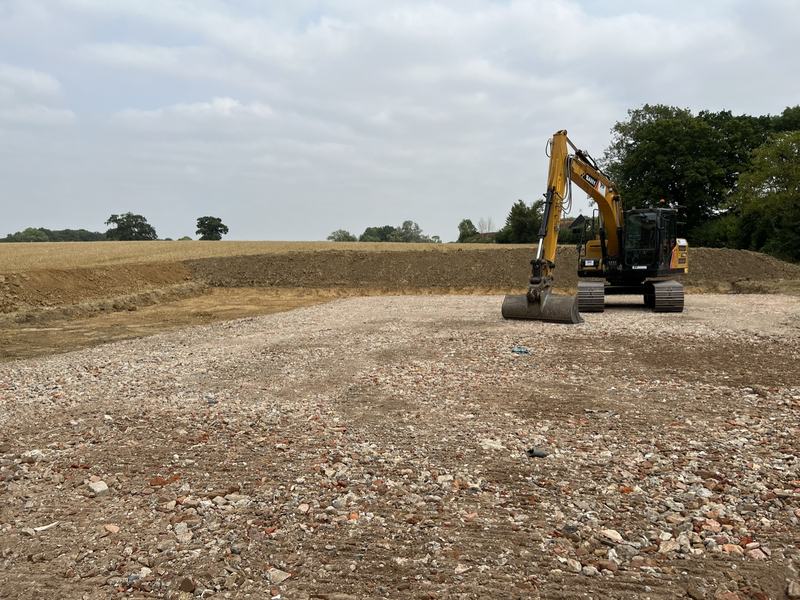 Large crushed hardcore hardstanding area with a tracked excavator and a field behind