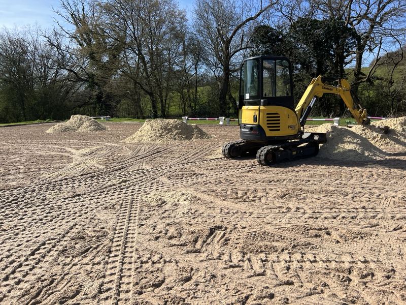 Yellow Yanmar mini digger beside heaps of new waxed silica sand mix on an outdoor arena, with trees behind