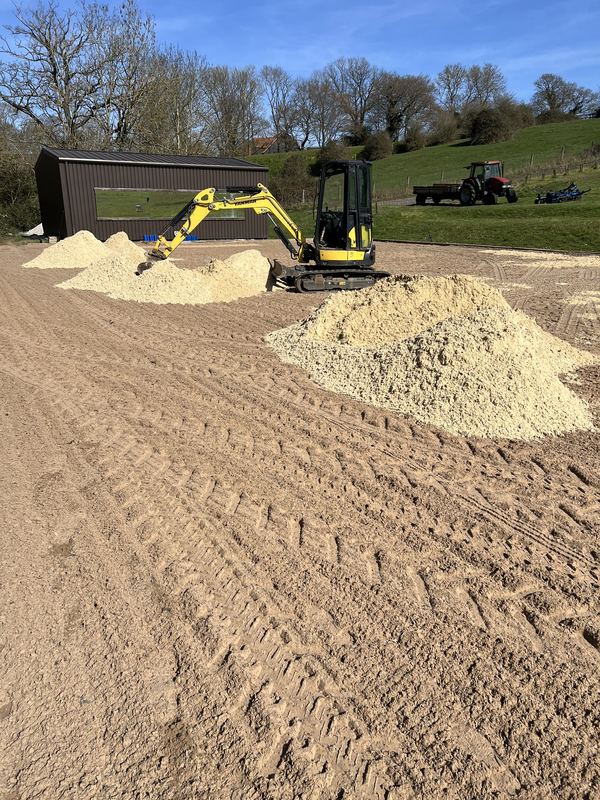Mini digger spreading heaps of new waxed silica sand mix across a ménage, with a timber barn and a red tractor behind