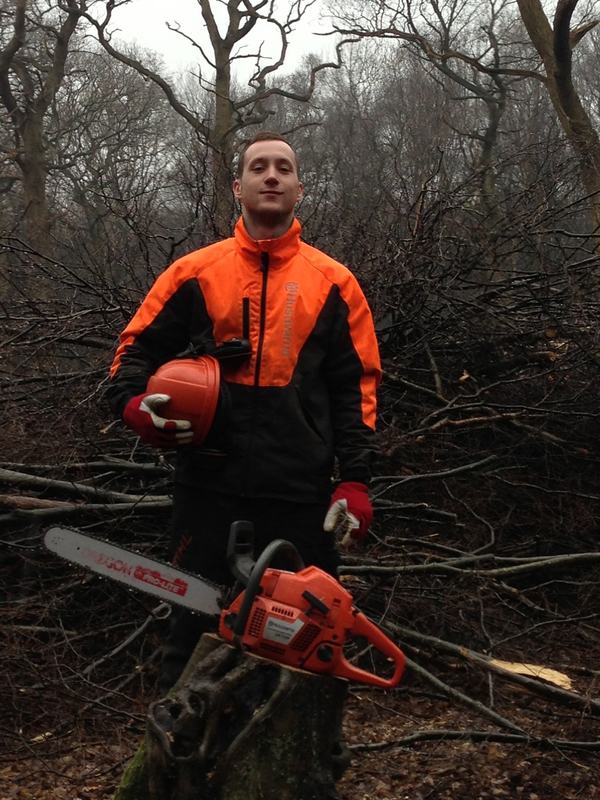Owner of South East Services Ltd in chainsaw safety gear, holding a helmet beside a chainsaw on a tree stump in woodland