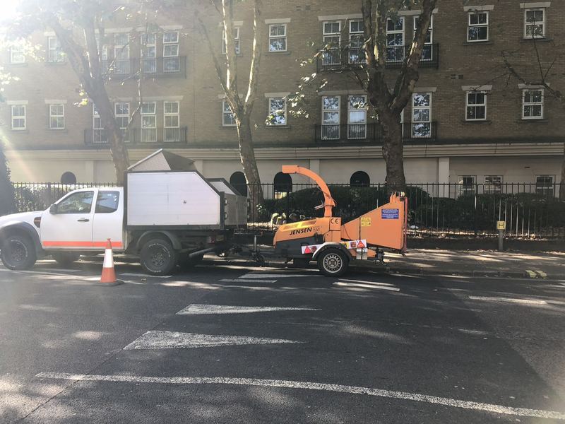Pickup truck with a tipper body towing a Jensen wood chipper, parked beside a tree-lined pavement