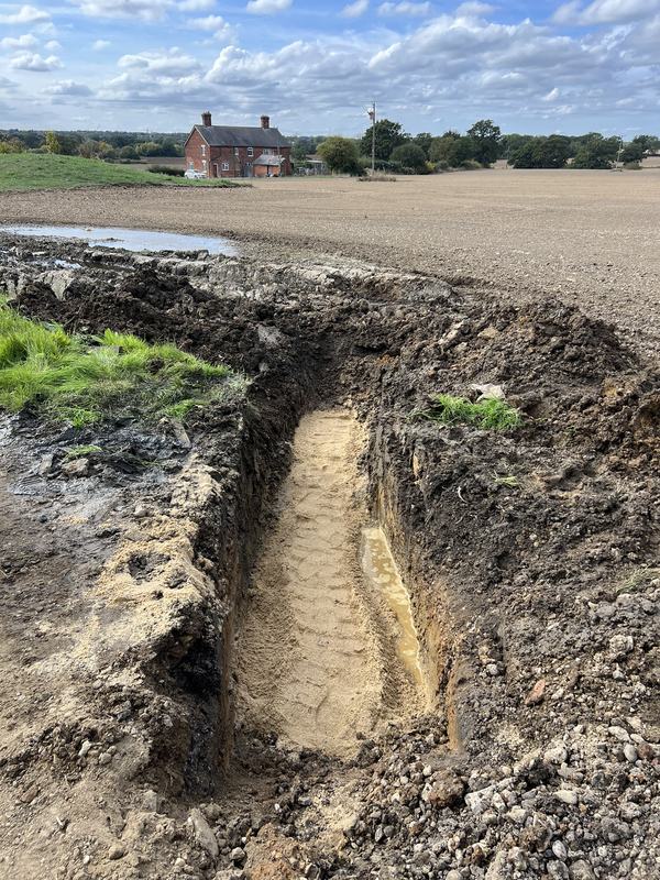 Long trench dug along a field edge with sand bedding in the base, with a farmhouse in the distance