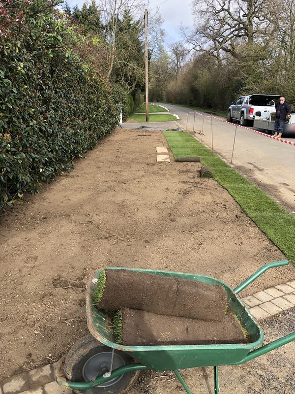Roadside verge stripped and levelled ready for turf, with turf rolls in a wheelbarrow and new turf being laid along the kerb